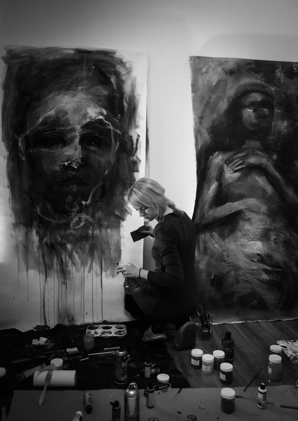 The artist working at the base of a portrait, surrounded by painting materials.