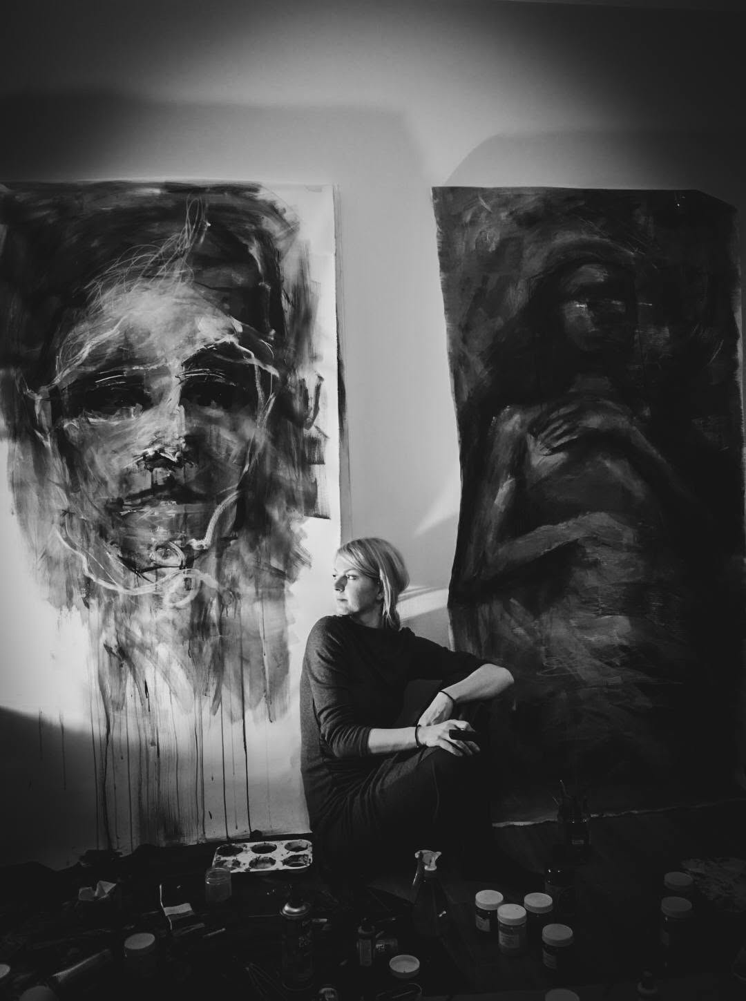 The artist seated between two figurative compositions in her studio.