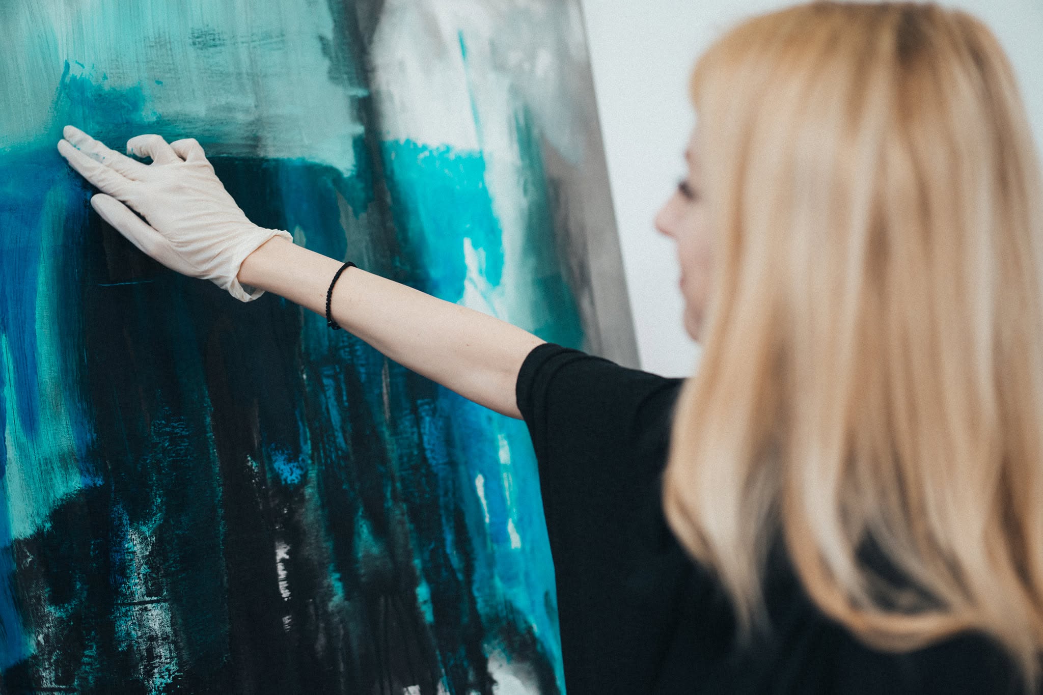 A close-up of the artist’s gloved hand working on a turquoise and black painted surface.