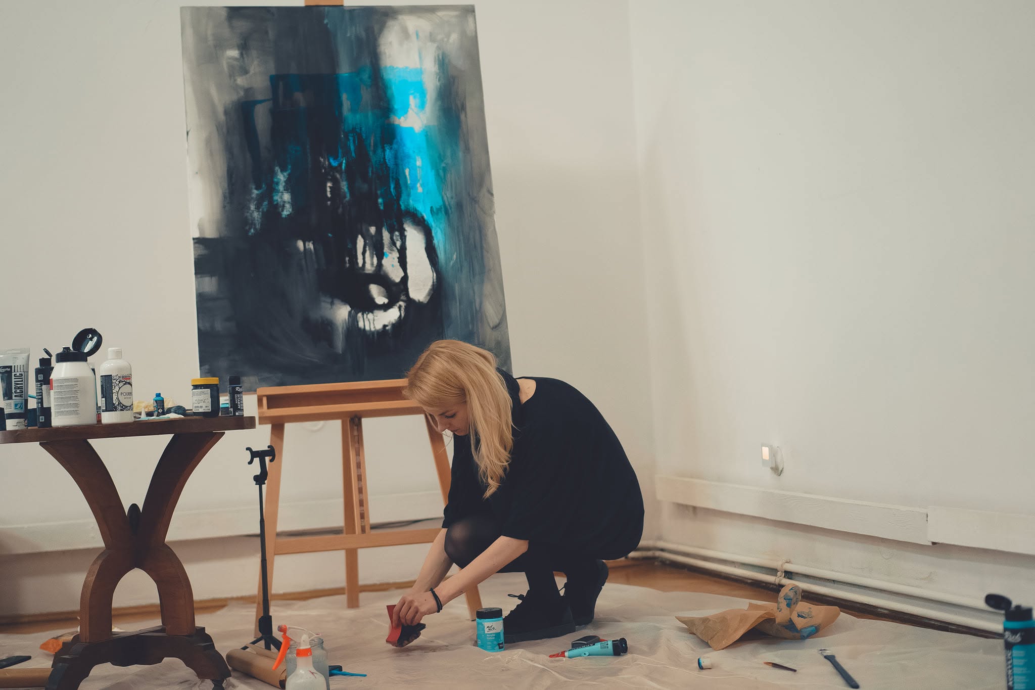 The artist preparing colour in front of the easel, with the canvas and painting materials in view.