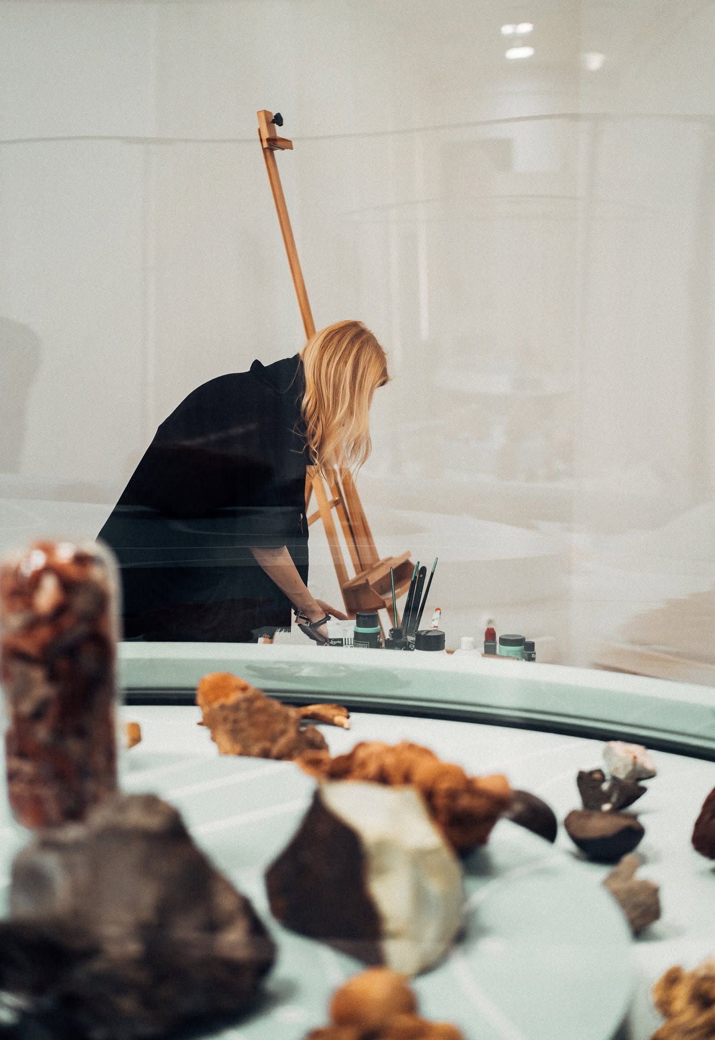 The artist preparing painting materials beside her easel, seen beyond a museum display case.